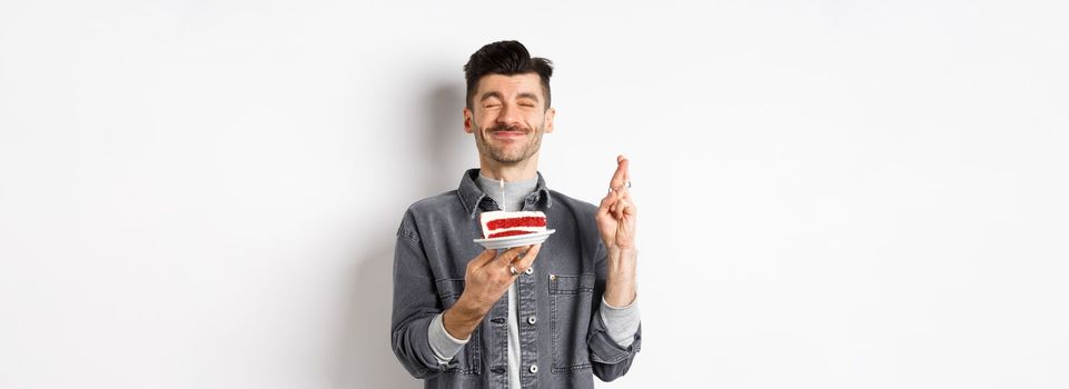 Hopeful birthday guy making wish with fingers crossed, holding bday cake on party, standing against white background.
