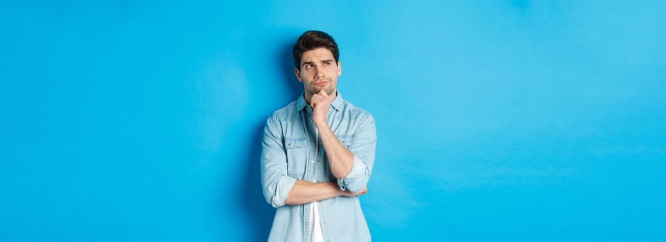 Image of adult bearded man 25s years, thinking about something, looking at upper left corner and pondering ideas, standing over blue background.