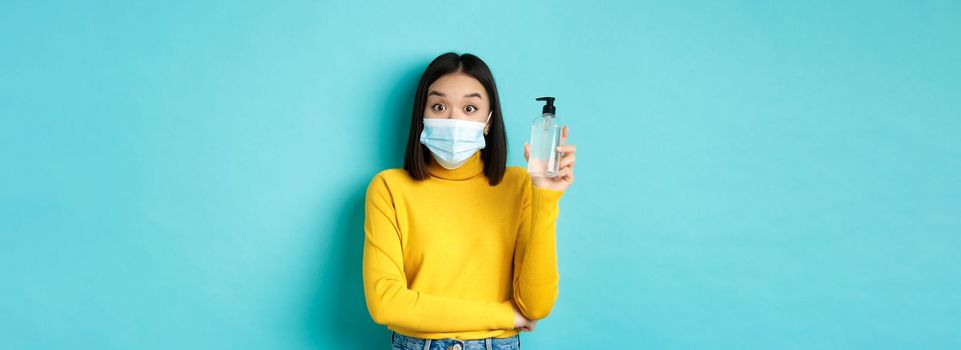 Covid-19, social distancing and pandemic concept. Cheerful asian woman showing hand sanitizer, recommend use antiseptic from coronavirus, wearing medical mask.