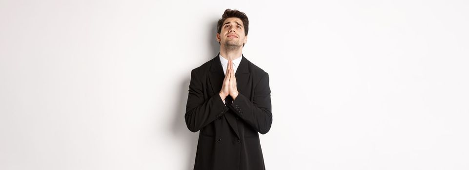 Troubled and hopeful man in black suit begging god, pleading and looking up, need help, standing over white background.