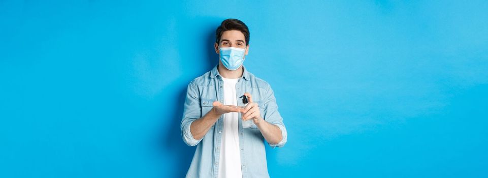 Concept of covid-19, pandemic and social distancing. Handsome young man in medical mask disinfecting hands with sanitizer, using antiseptic, standing against blue background.