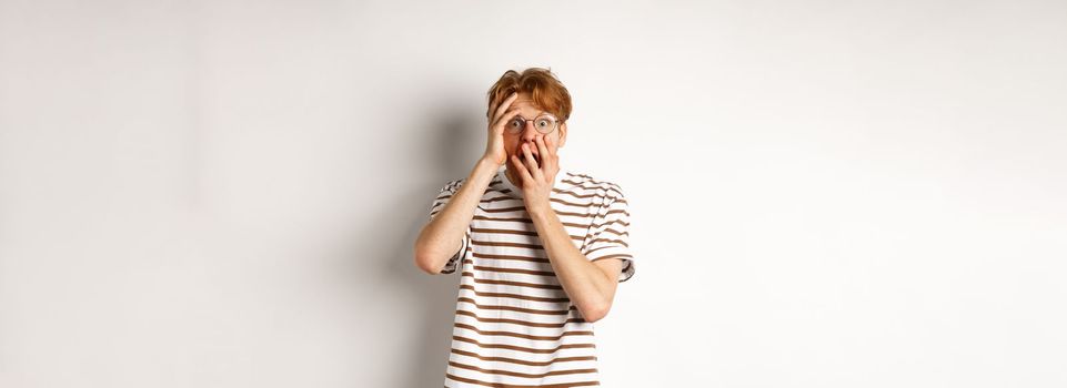 Scared redhead guy screaming terrified, holding hands on face from fear, standing over white background.