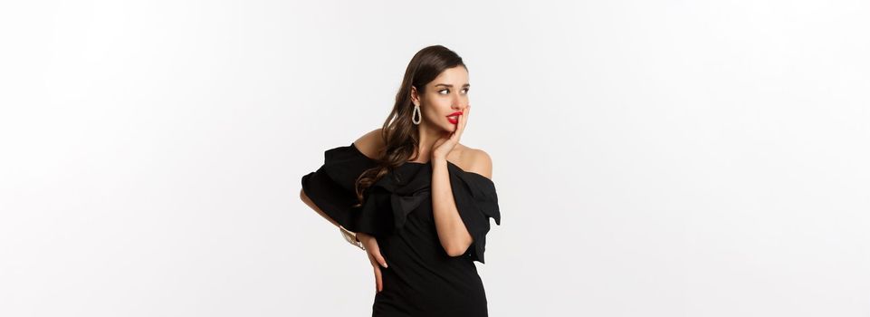 Fashion and beauty. Image of stylish beautiful woman in black dress and makeup, looking left with temptation, touching red lips, standing over white background.