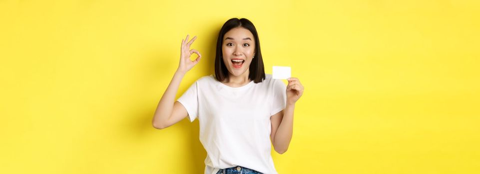 Young asian woman in casual white t-shirt showing plastic credit card and okay gesture, recommend bank, smiling at camera, yellow background.