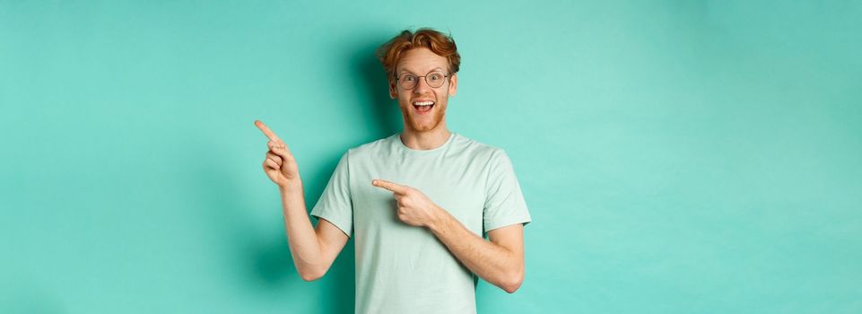 Handsome smiling man with red hair and beard, looking amused and pointing at upper right corner, showing promo offer, standing over mint background.