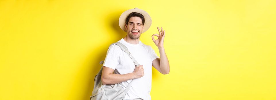 Tourism, travelling and holidays concept. Happy tourist going on vacation, holding backpack and showing okay sign smiling satisfied, standing against yellow background.