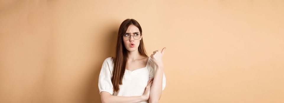 Confused and suspicious young woman in glasses frowning, pointing and looking aside with displeased face expression, standing on beige background.