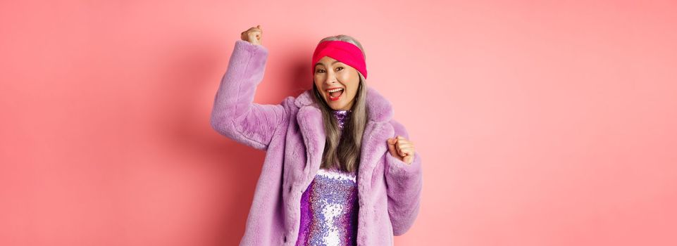 Shopping concept. Cheerful senior asian woman dancing and having fun, wearing stylish purple faux fur coat and dress, winning prize and smiling, standing over pink background.