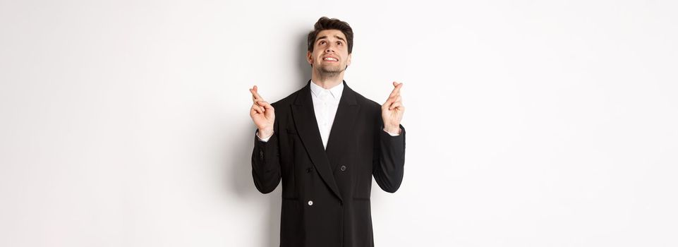 Portrait of tensed and worried handsome businessman, crossing fingers and looking up, begging god, making a wish, standing against white background in black suit.