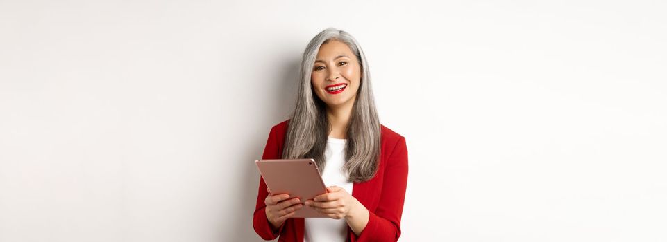 Business. Successful senior businesswoman working with digital tablet and smiling, standing in red blazer over white background.