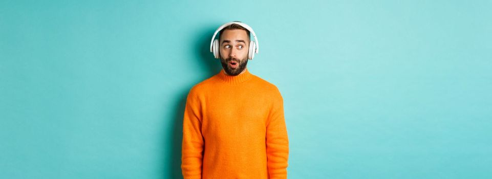 Amazed adult man listening music in headphones, looking at camera impressed with sound, standing over turquoise background.