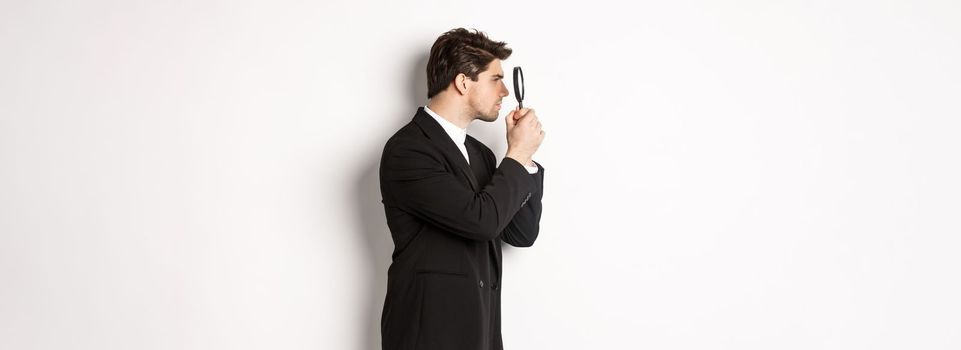 Profile shot of handsome businessman in black suit, looking through magnifying glass and searching for something, standing over white background.
