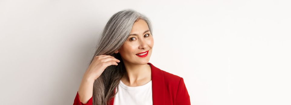 Beauty and haircare concept. Close up of elegant asian senior woman showing shiny and healthy grey hair, smiling and looking aside, white background.