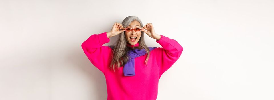 Fashionable asian senior woman trying new sunglasses, looking amazed and cheerful at camera, standing in trendy sweater against white background.