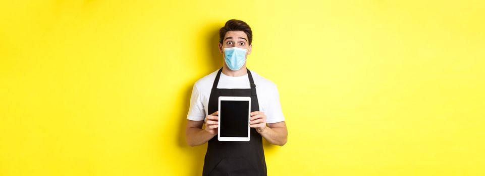Concept of covid-19, small business and pandemic. Surprised waiter in black apron and medical mask showing tablet screen, standing over yellow background.