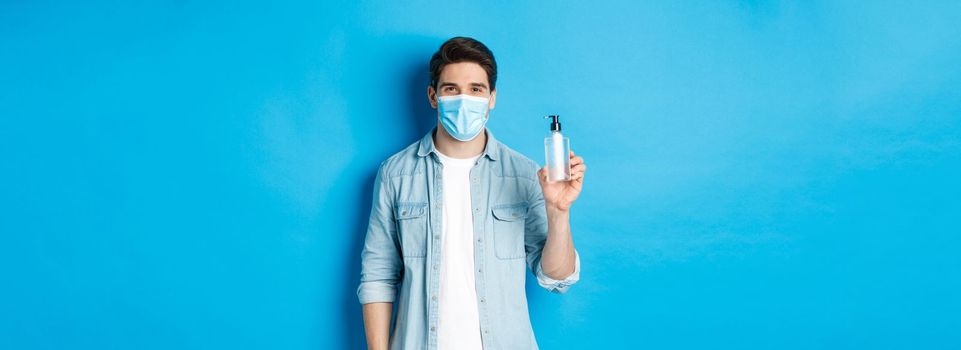 Concept of covid-19, pandemic and social distancing. Young man in medical mask showing hand sanitizer, standing against blue background.