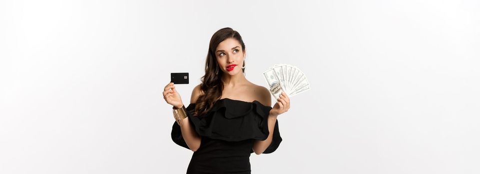 Fashion and shopping concept. Elegant woman in black dress, red lipstick, holding money and credit card, making choice, thinking, standing over white background.