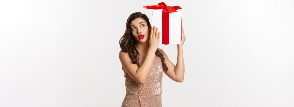 Holidays, celebration concept. Beautiful woman in luxury dress trying to guess what inside gift box, shaking Christmas present with curious face, white background.