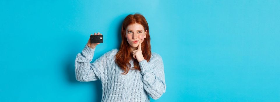 Image of thoughtful redhead girl thinking about shopping, showing credit card and pondering, standing over blue background.