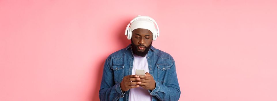 Man looking serious while reading messagin on phone, listening music in headphones, standing over pink background.