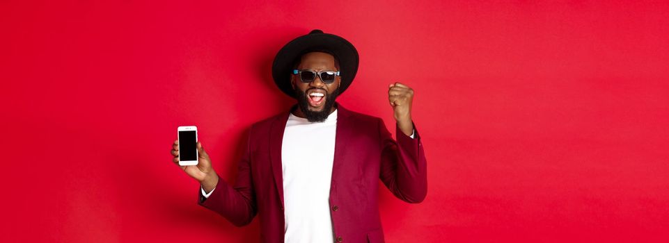 Cheerful winning Black man showing smartphone screen, rejoicing and demonstrating application, standing over red background.