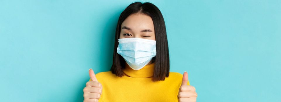 Covid-19, social distancing and pandemic concept. Cute asian girl in medical mask winking at camera, showing thumbs up, good job gesture, praise nice work, blue background.