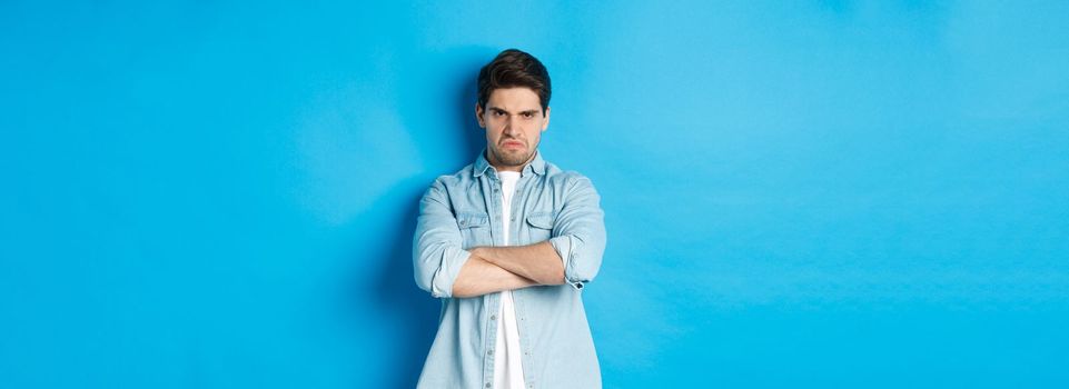 Angry bearded guy crossing arms on chest, frowning, looking mad and offended, standing against blue background.