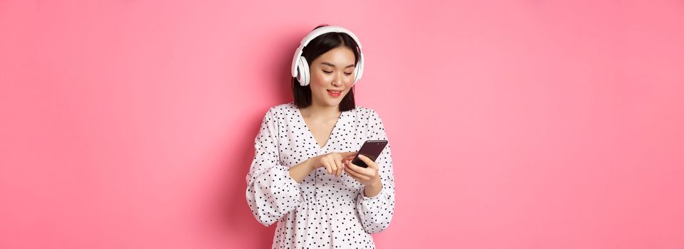 Beautiful asian woman texting message on smartphone, listening music in headphones, standing over pink background.