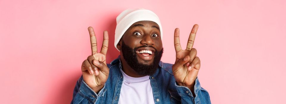 Close-up of cheerful african-american hipster in beanie smiling, showing peace signs, standing happy against pink background.