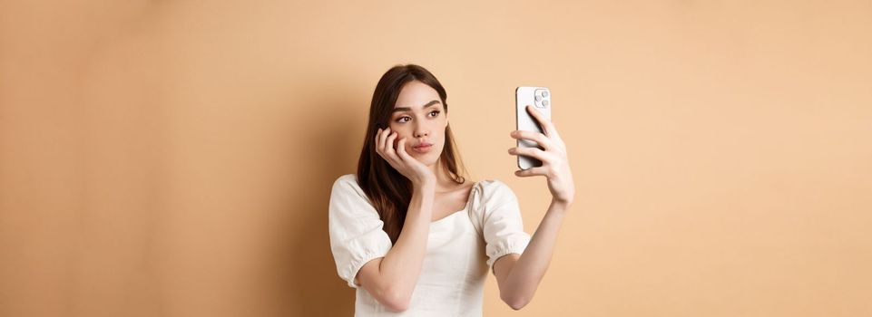 Stylish girl taking selfie, posing for mobile camera with photo filters, making cute face, standing on beige background.