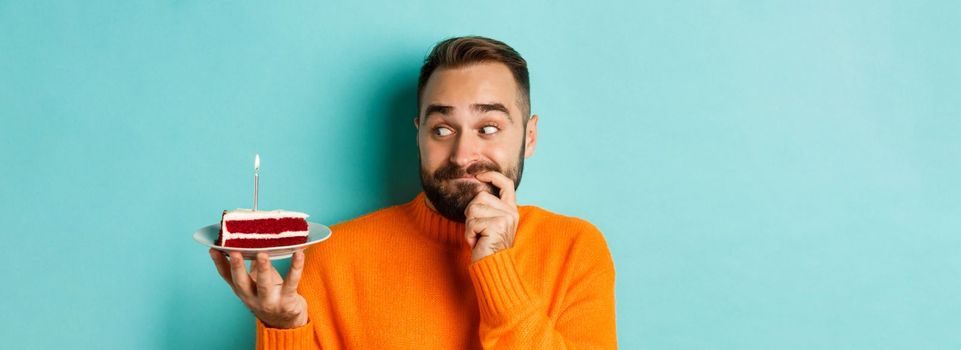 Close-up of happy adult man celebrating birthday, holding bday cake with candle and making wish, standing against turquoise background.