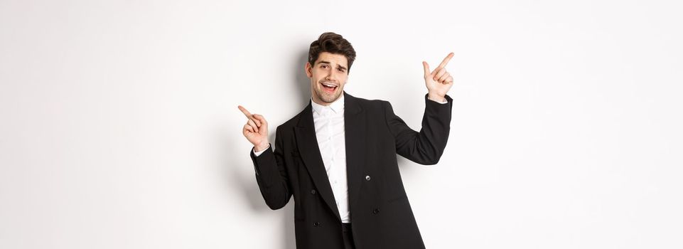 Handsome guy dancing and partying in black suit, pointing fingers sideways, showing two promo banners, standing against white background.