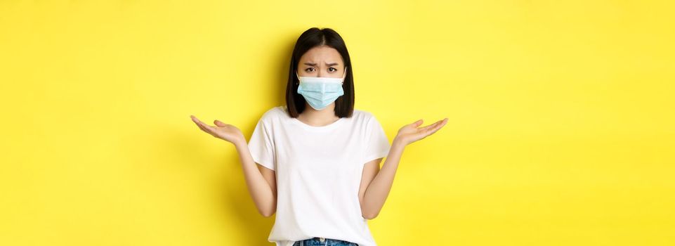 Covid-19, quarantine and social distancing concept. Confused asian woman in medical mask shrugging shoulders, spread hands sideways clueless, know nothing, yellow background.
