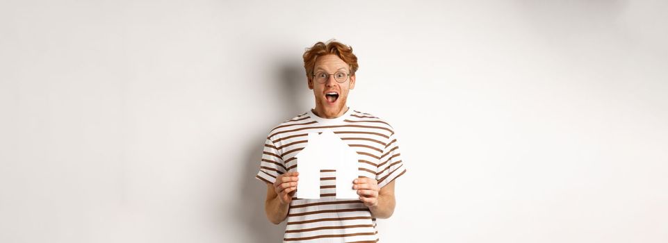 Happy and surprised redhead man winning house, holding paper home model and staring at camera, standing joyful over white background.