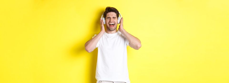 Happy guy listening music in new headphones, buying earphones on black friday, standing over yellow background.