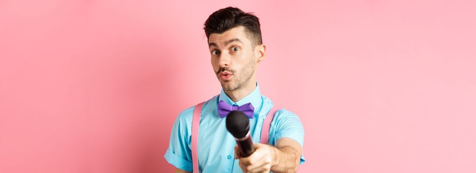 Handsome male show host stretch out hand with microphone, asking for interview or comments, standing on pink background.