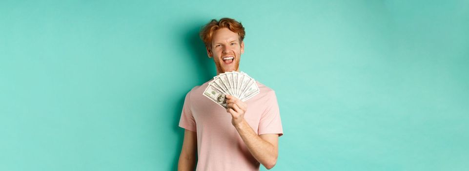 Happy redhead man in t-shirt showing money in dollars and smiling, making smug faces after winning cash, standing over turquoise background.