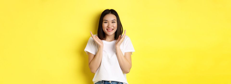 Beauty and fashion concept. Excited asian woman clap hands and smiling happy at camera, standing in white t-shirt against yellow background.