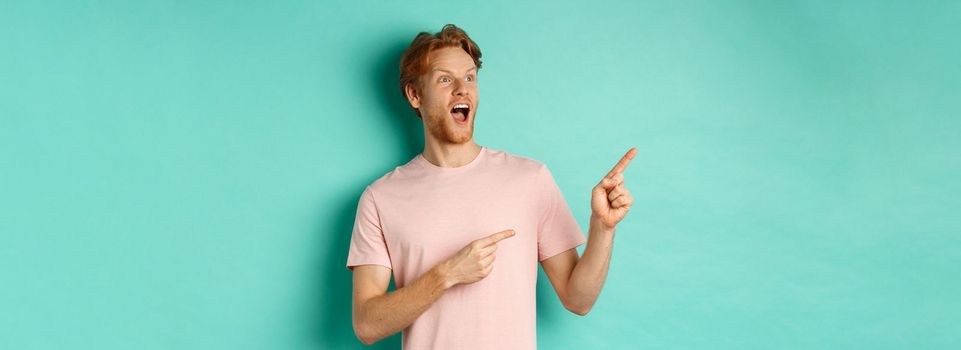 Amazed redhead man in t-shirt checking out promo, gasping in awe and pointing fingers at upper left corner, standing over mint background.