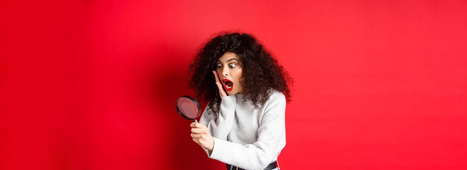 Shocked girl look through magnifying glass with dropped jaw, seeing something amazing, standing on red background.