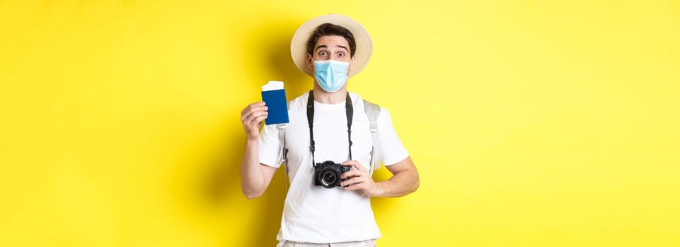 Concept of covid-19, travelling and quarantine. Happy man tourist with camera, showing passport and tickets for vacation, going on trip during pandemic, yellow background.