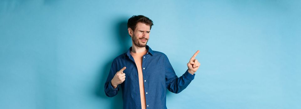 Skeptical young man pointing and looking left with disappointment, cringe from something bad, standing upset on blue background.