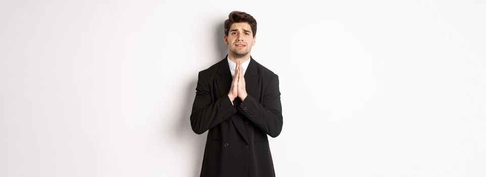 Image of handsome businessman in suit need help, begging you, holding hands in plead gesture and looking desperate, standing over white background.