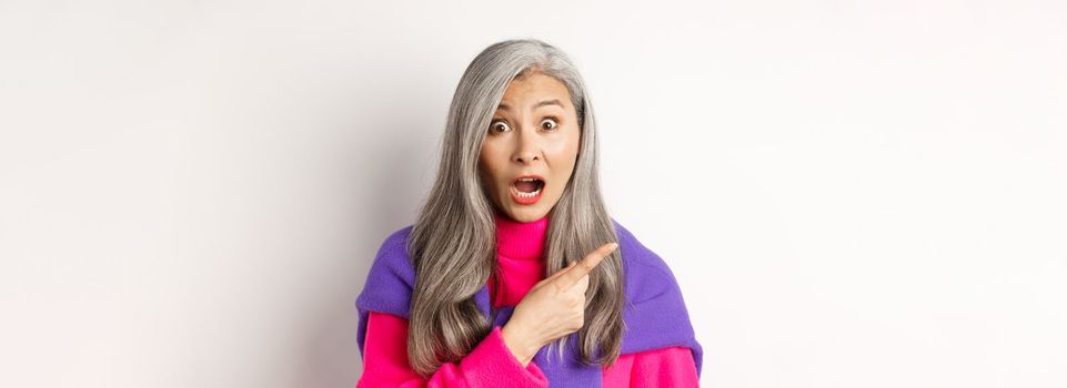 Close-up of amazed and intrigued asian senior woman pointing finger left at something cool, staring at camera impressed, standing over white background.