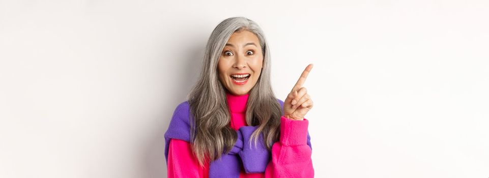 Close up of beautiful asian female model pointing finger at upper left corner, smiling and looking happy at camera, showing promo deal, white background.