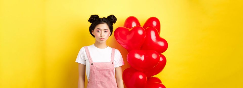 Valentines day concept. Sad and gloomy asian woman in makeup, frowning upset, standing near red hearts balloons and feeling upset, standing on yellow background.