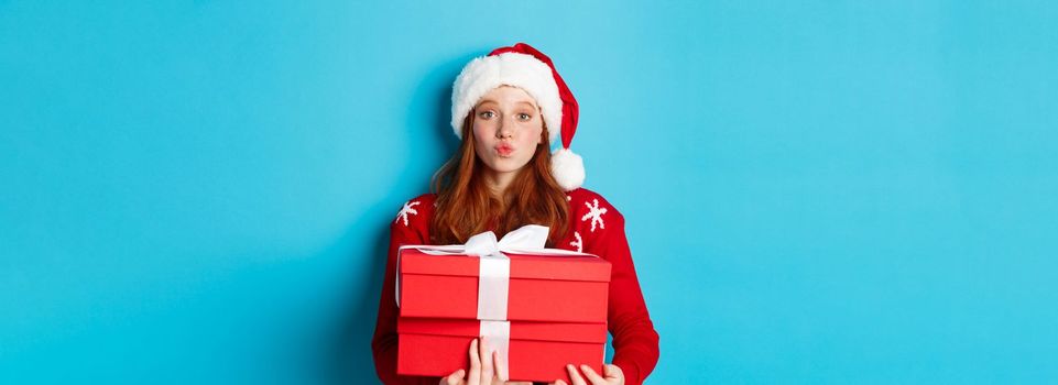 Happy holidays and Christmas concept. Cute redhead girl holding presents and pucker lips for kiss, wearing santa hat and funny sweater, blue background.