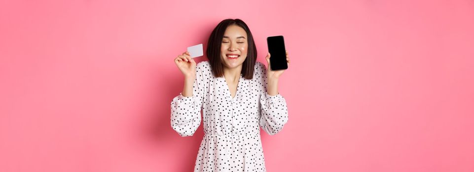 Joyful korean woman showing smartphone screen and credit card, paying for internet order, demonstrating online shopping app, standing over pink background.