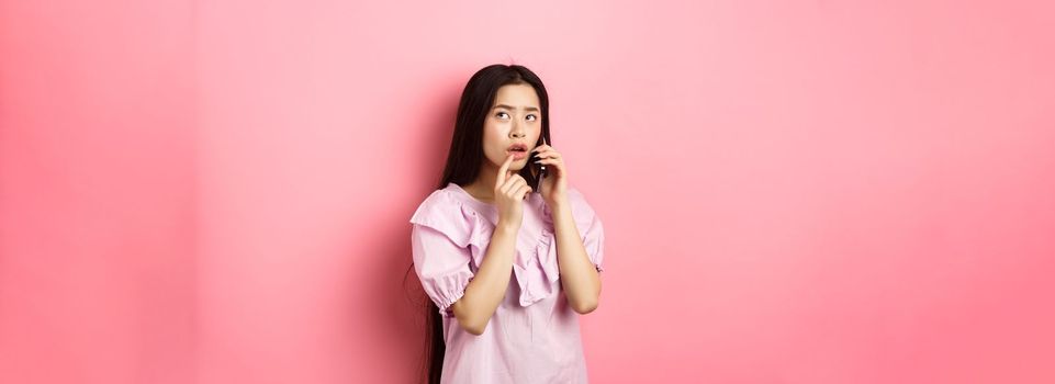 Thoughtful asian woman talking on mobile phone and thinking, touching lip and look up pensive, standing against pink background.