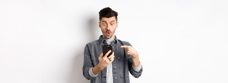 Amazed young man showing cool thing on smartphone, looking and pointing at phone impressed, standing against white background.